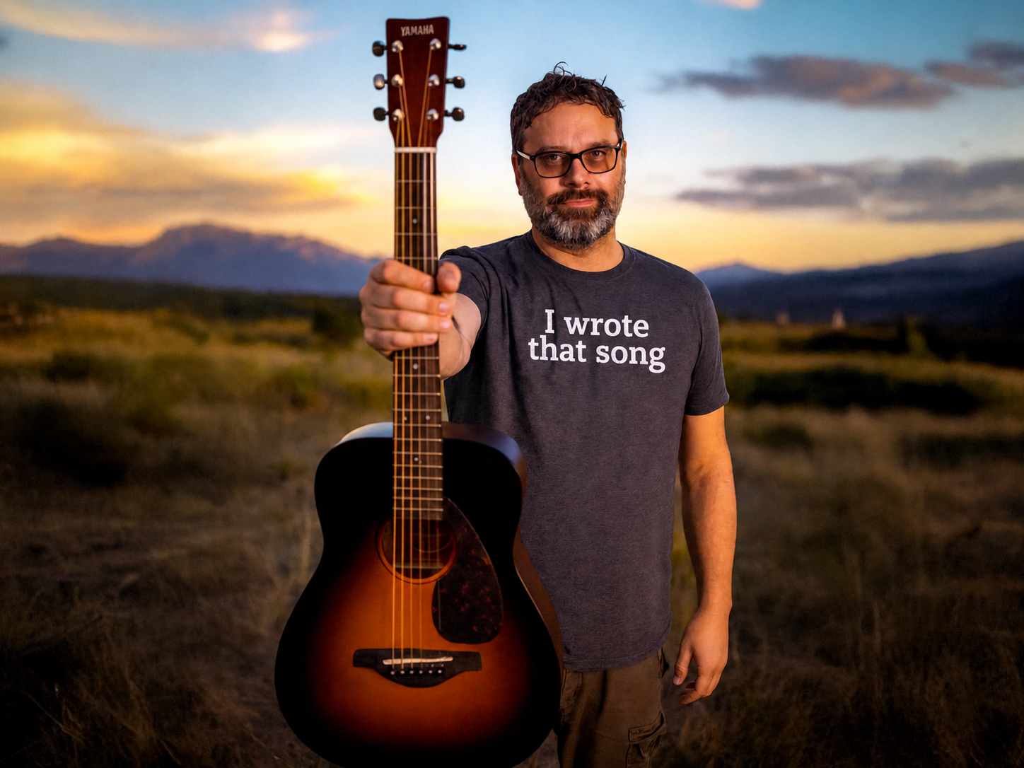 Galen Marten holding a guitar toward the camera at sunset, wearing an I wrote that song T-shirt